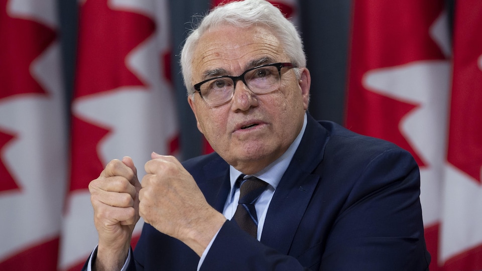 Raymond Theberg, seated in front of the Canadian flags, answers a question with my fist closed.